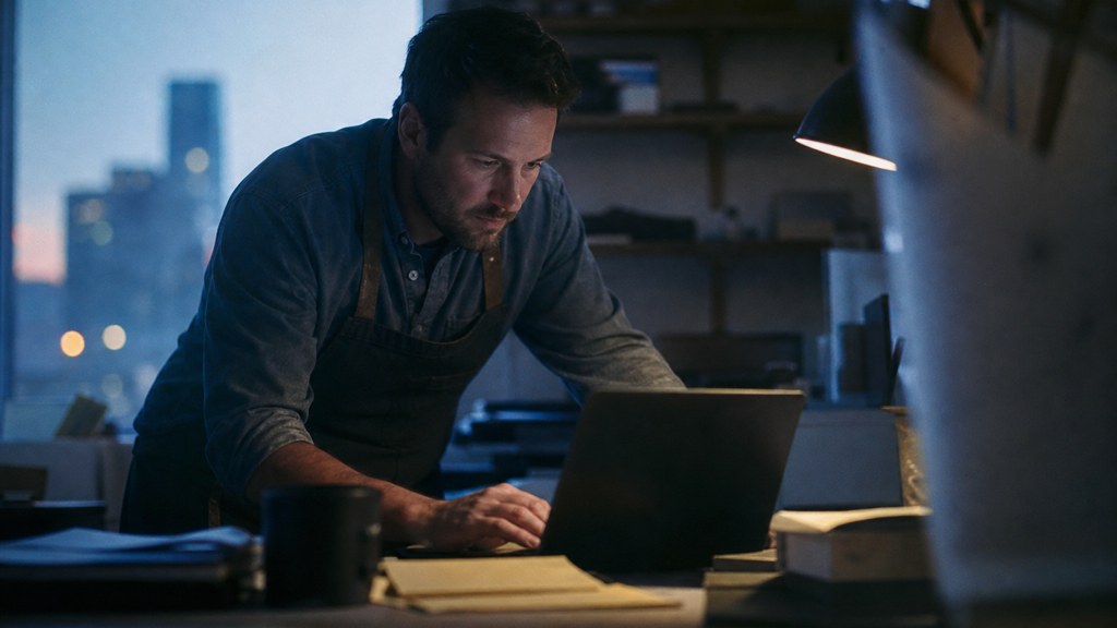 A small-business owner in an apron leaning over a laptop on a cluttered back-office desk late in the day, photographed in Oklahoma City