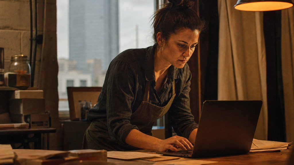A small-business owner in an apron leaning over a laptop on a cluttered back-office desk late in the day, photographed in Oklahoma City