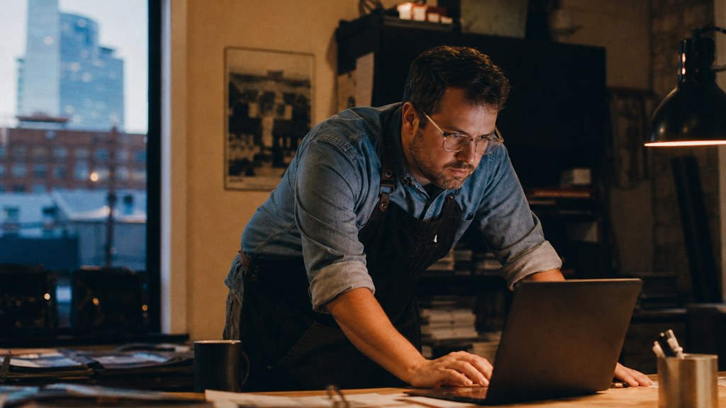 A small-business owner in an apron leaning over a laptop on a cluttered back-office desk late in the day, photographed in Oklahoma City