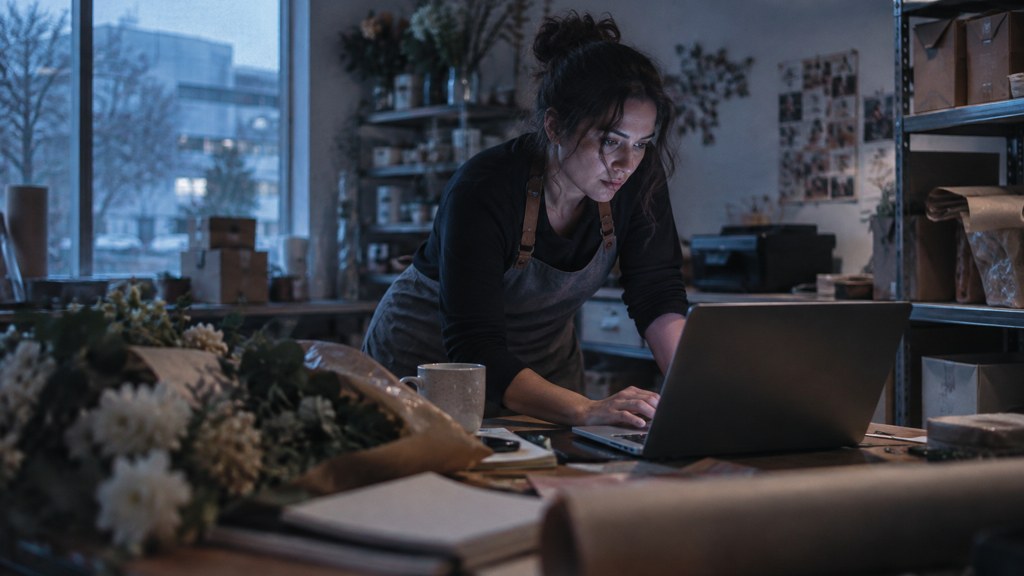A small-business owner in an apron leaning over a laptop on a cluttered back-office desk late in the day, photographed in Plano
