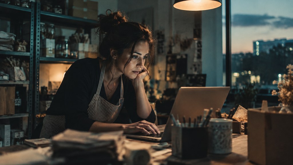 A small-business owner in an apron leaning over a laptop on a cluttered back-office desk late in the day, photographed in Plano