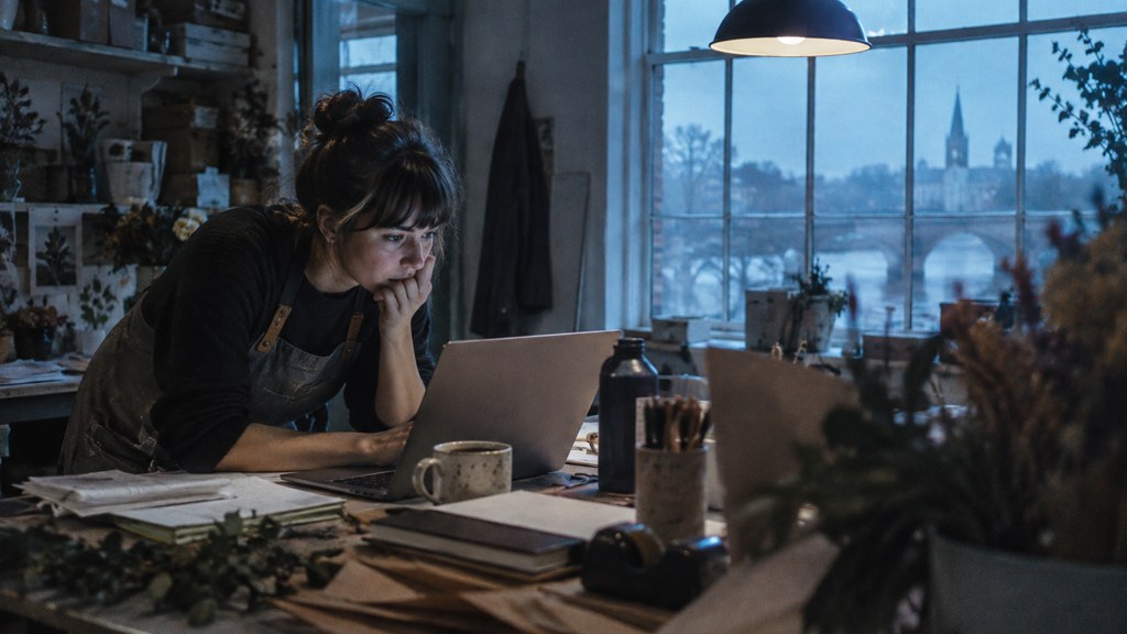 A small-business owner in an apron leaning over a laptop on a cluttered back-office desk late in the day, photographed in Richmond