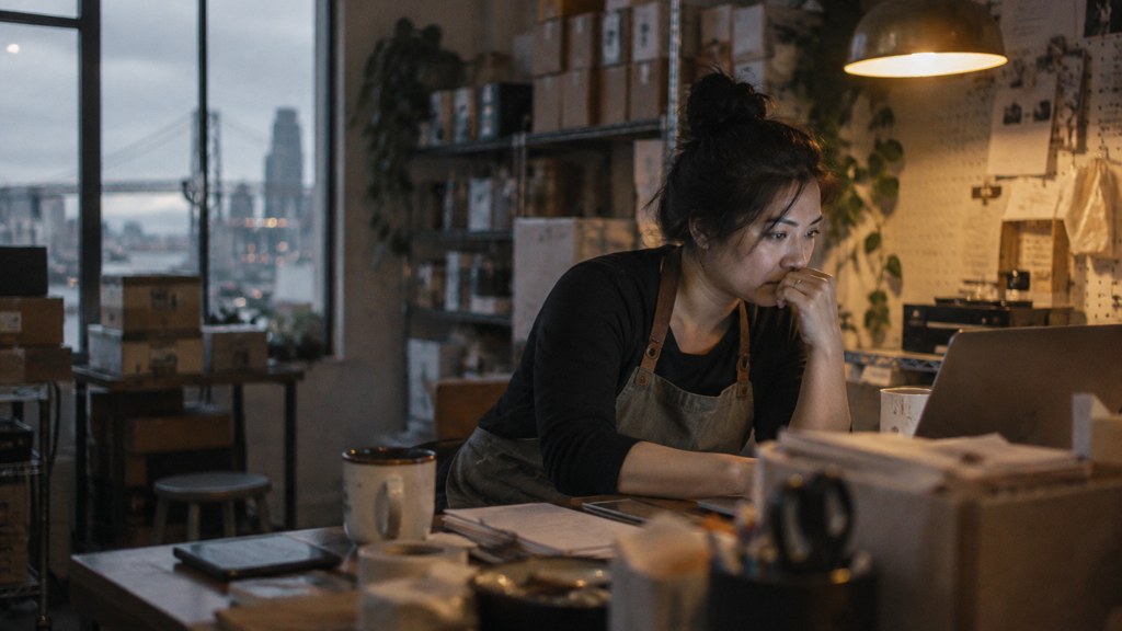 A small-business owner in an apron leaning over a laptop on a cluttered back-office desk late in the day, photographed in San Francisco