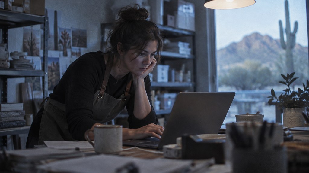 A small-business owner in an apron leaning over a laptop on a cluttered back-office desk late in the day, photographed in Scottsdale