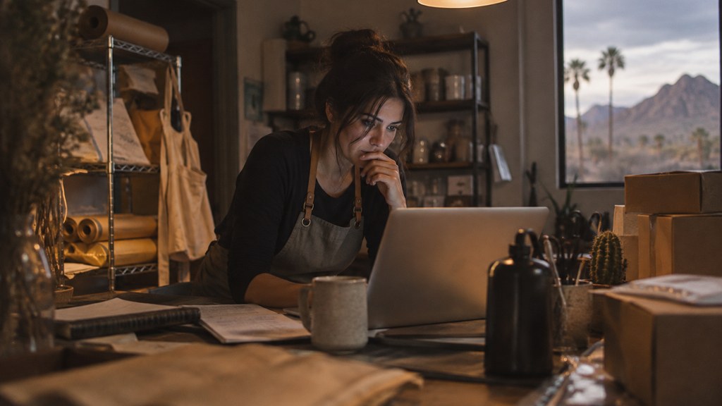 A small-business owner in an apron leaning over a laptop on a cluttered back-office desk late in the day, photographed in Scottsdale