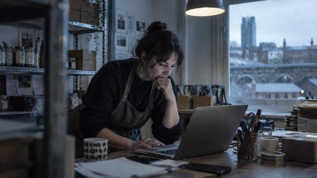 A small-business owner in an apron leaning over a laptop on a cluttered back-office desk late in the day, photographed in Sheffield