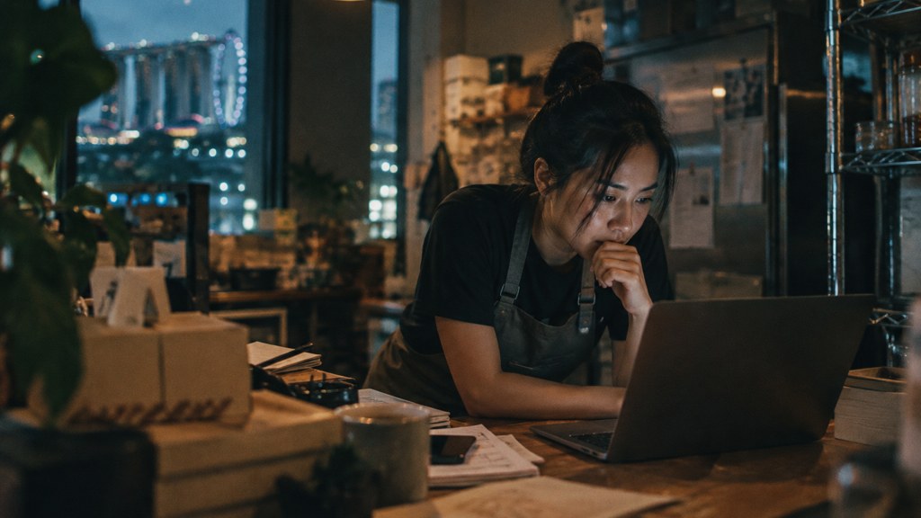 A small-business owner in an apron leaning over a laptop on a cluttered back-office desk late in the day, photographed in Singapore