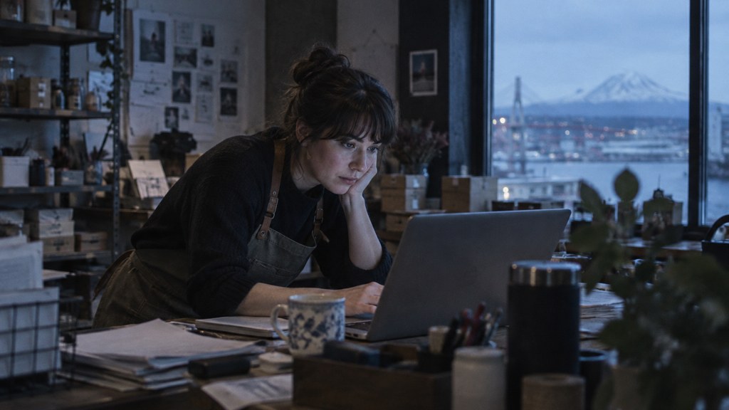 A small-business owner in an apron leaning over a laptop on a cluttered back-office desk late in the day, photographed in Tacoma