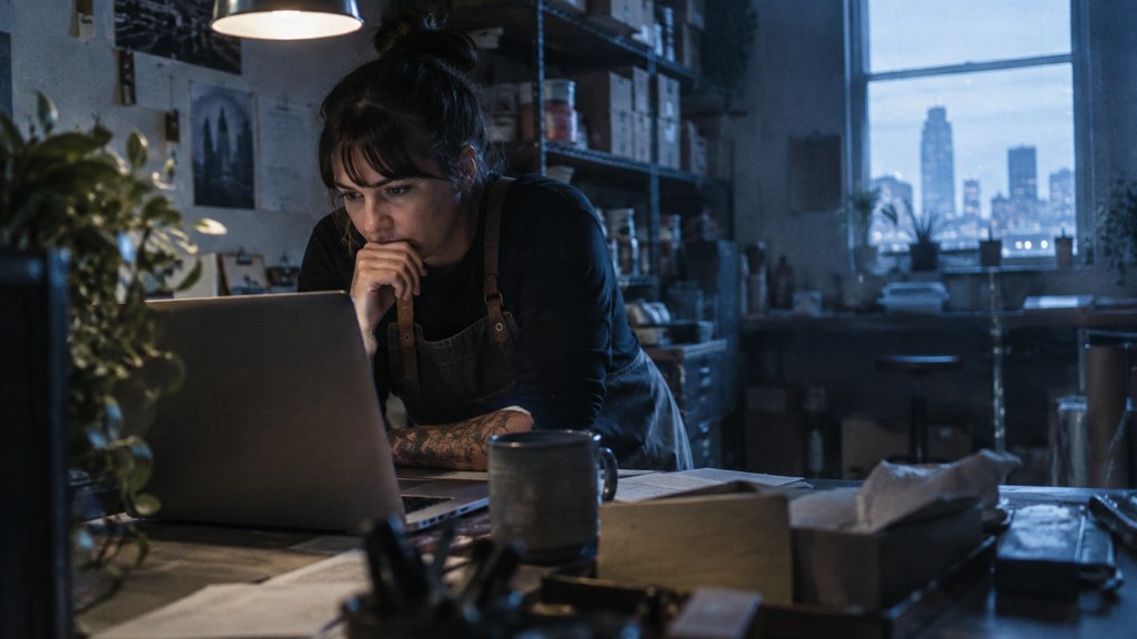 A small-business owner in an apron leaning over a laptop on a cluttered back-office desk late in the day, photographed in Tulsa