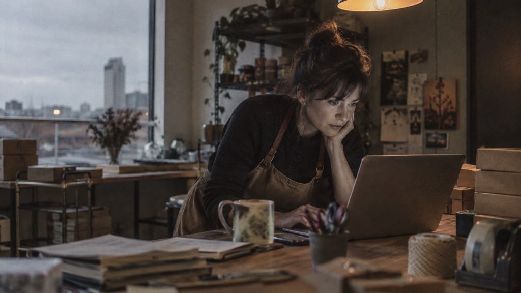 A small-business owner in an apron leaning over a laptop on a cluttered back-office desk late in the day, photographed in Wichita
