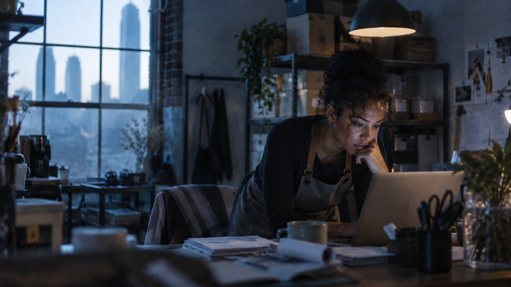 A small-business owner in an apron leaning over a laptop on a cluttered back-office desk late in the day, photographed in Atlanta