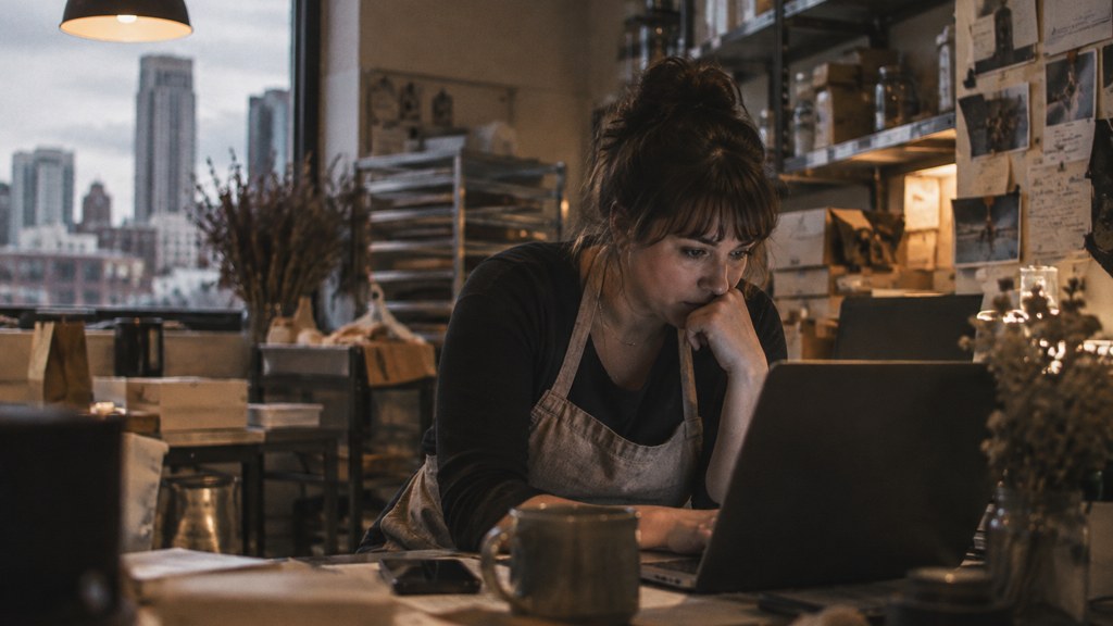 A small-business owner in an apron leaning over a laptop on a cluttered back-office desk late in the day, photographed in Denver