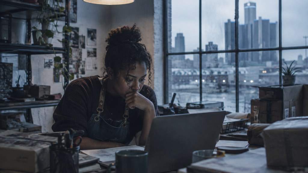 A small-business owner in an apron leaning over a laptop on a cluttered back-office desk late in the day, photographed in Detroit