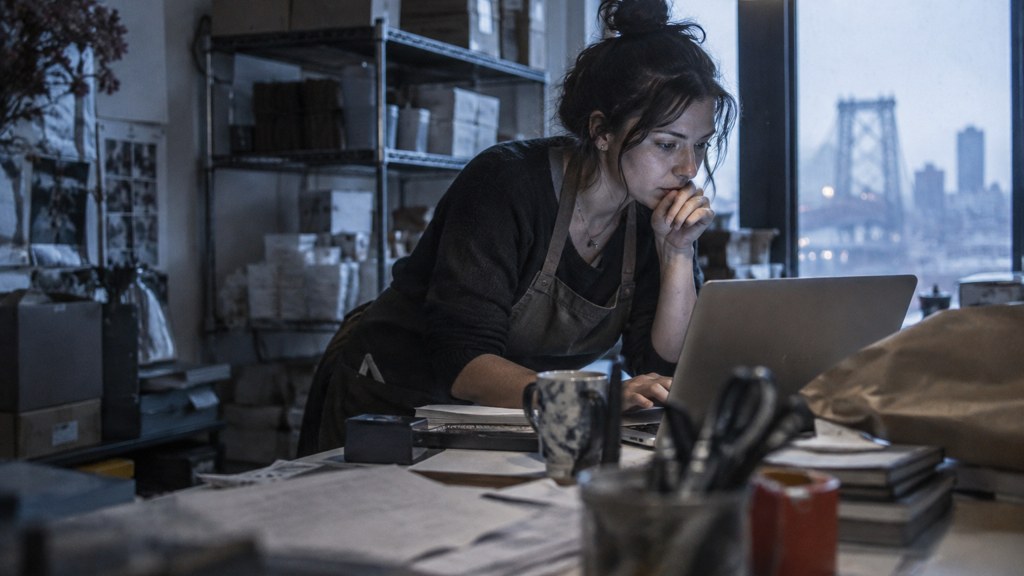 A small-business owner in an apron leaning over a laptop on a cluttered back-office desk late in the day, photographed in New York