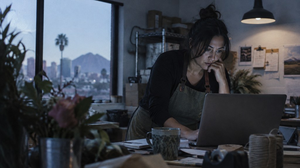 A small-business owner in an apron leaning over a laptop on a cluttered back-office desk late in the day, photographed in Phoenix