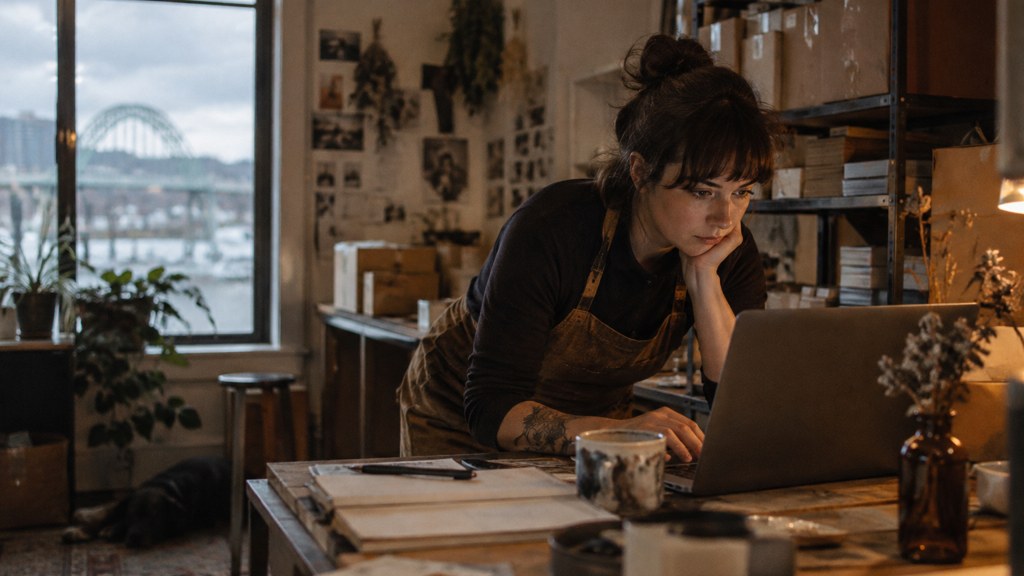 A small-business owner in an apron leaning over a laptop on a cluttered back-office desk late in the day, photographed in Portland