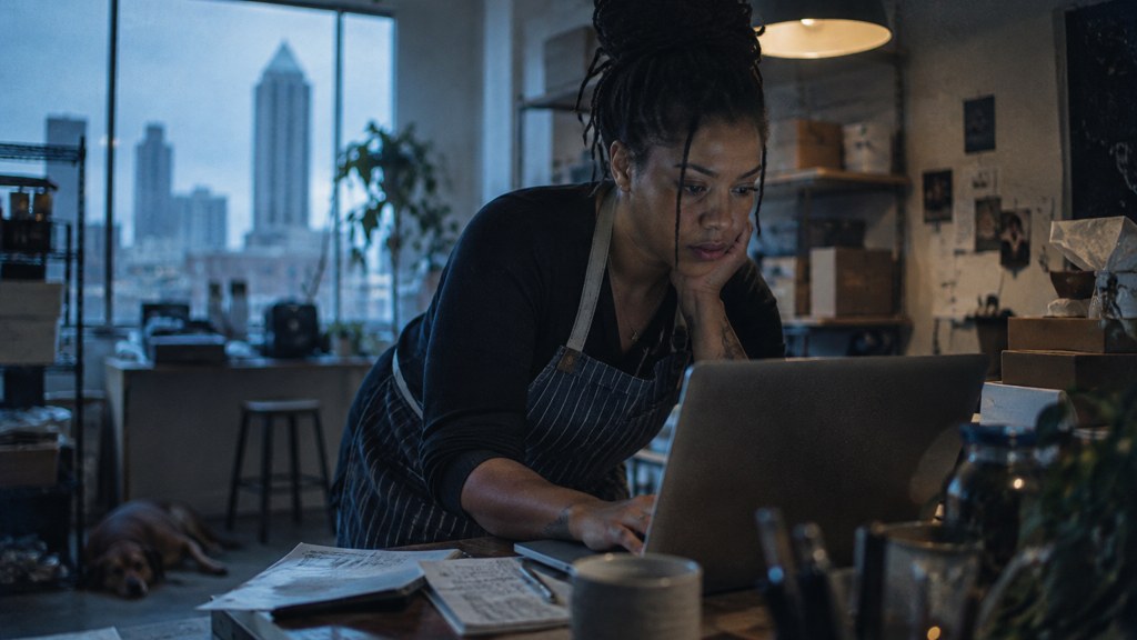 A small-business owner in an apron leaning over a laptop on a cluttered back-office desk late in the day, photographed in Atlanta