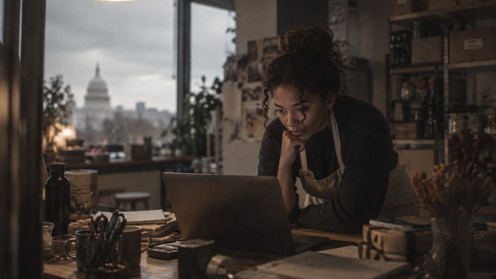 A small-business owner in an apron leaning over a laptop on a cluttered back-office desk late in the day, photographed in Washington DC