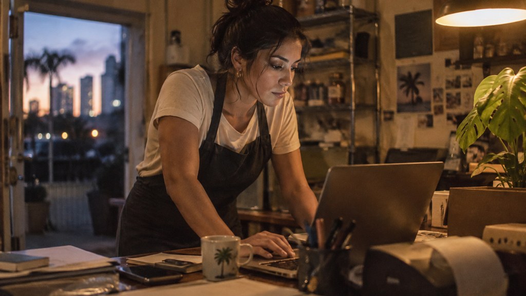 A small-business owner in an apron leaning over a laptop on a cluttered back-office desk late in the day, photographed in Miami