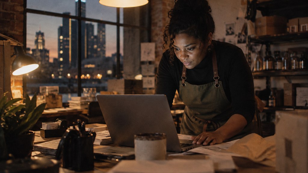 A small-business owner in an apron leaning over a laptop on a cluttered back-office desk late in the day, photographed in Detroit
