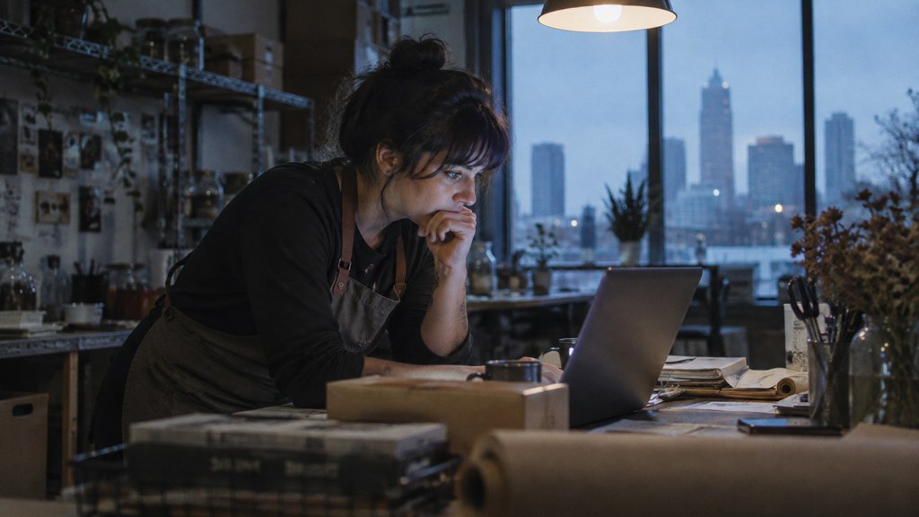 A small-business owner in an apron leaning over a laptop on a cluttered back-office desk late in the day, photographed in Charlotte