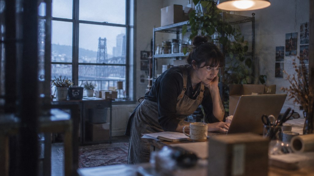 A small-business owner in an apron leaning over a laptop on a cluttered back-office desk late in the day, photographed in Portland