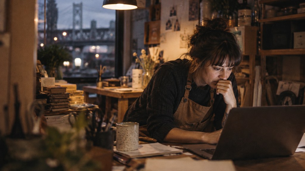 A small-business owner in an apron leaning over a laptop on a cluttered back-office desk late in the day, photographed in Portland