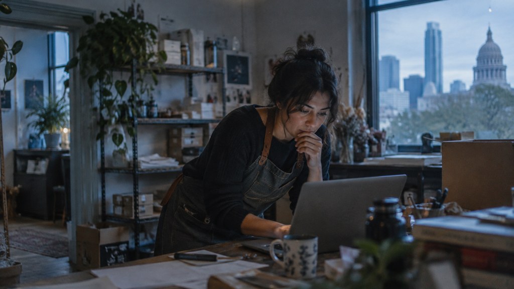 A small-business owner in an apron leaning over a laptop on a cluttered back-office desk late in the day, photographed in Austin