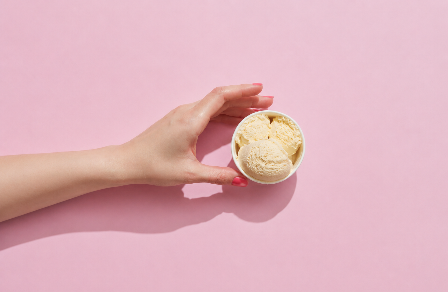 Hand with pink nail polish holding a cup with three scoops of vanilla ice cream on pink background.