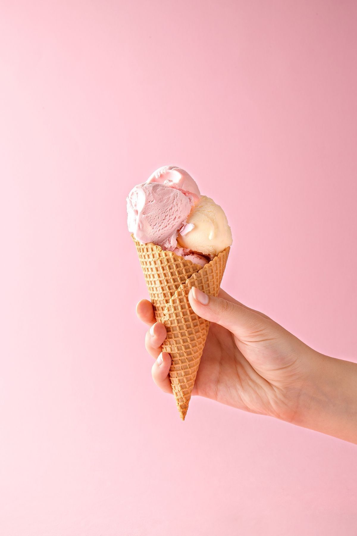 Hand holding a waffle cone with three scoops of ice cream in pink and cream colors against pink background.