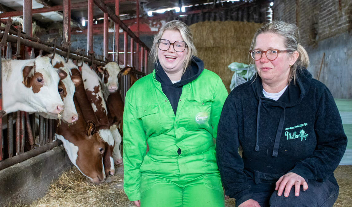 Two women sitting in a barn near feeding cows, one wearing a green coverall and the other a black hoodie.