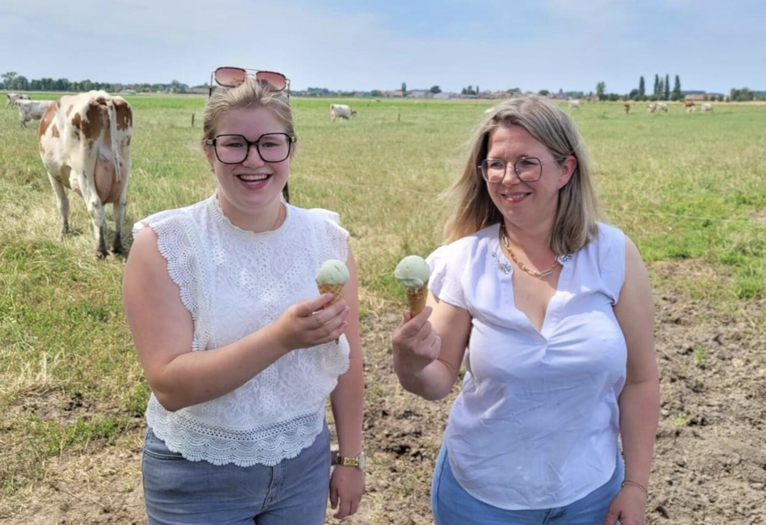Two women smiling and holding green ice cream cones in a grassy field with cows in the background.