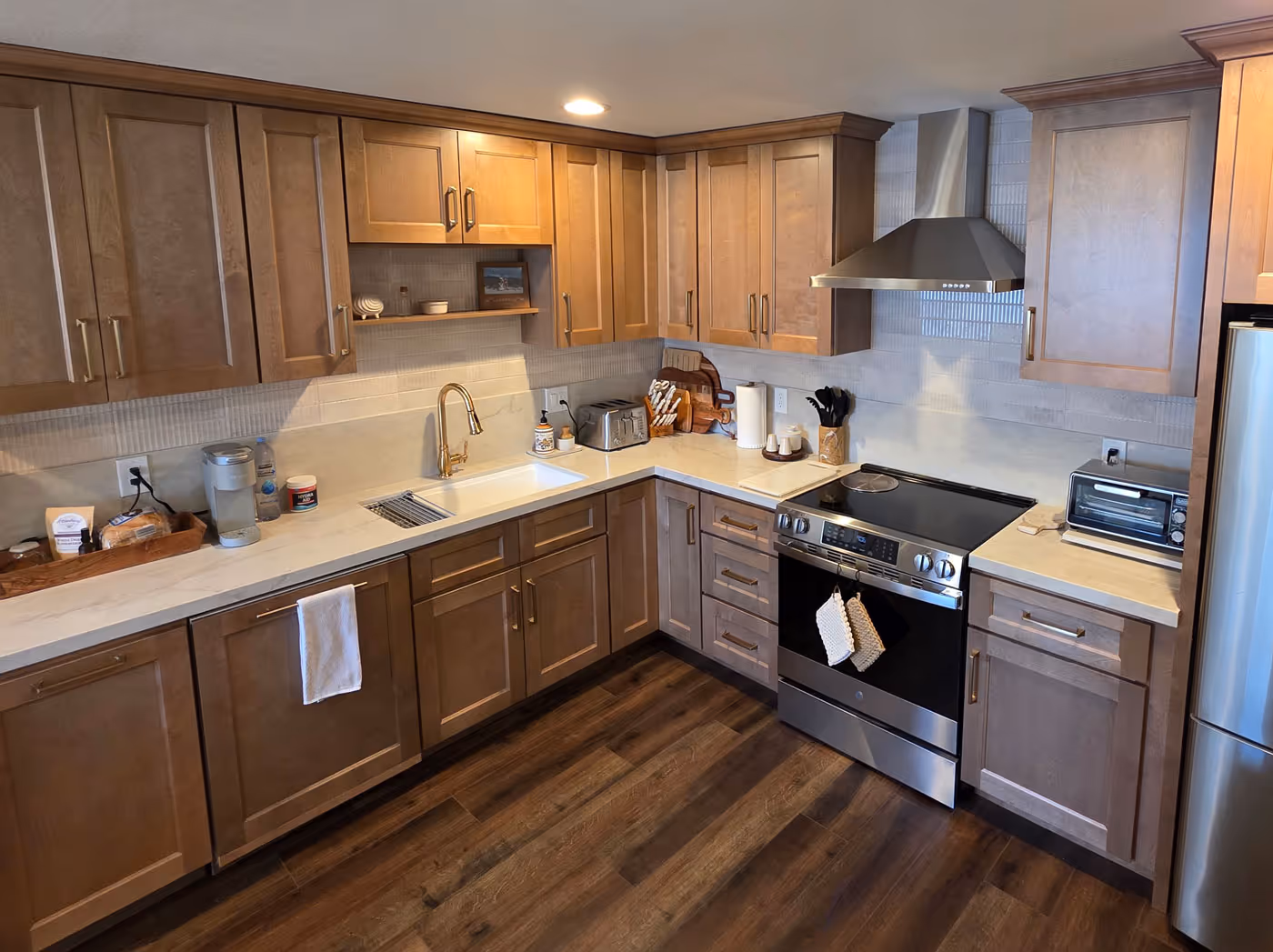 Modern kitchen with brown shaker cabinets, white countertops, stainless steel appliances, and wood flooring.