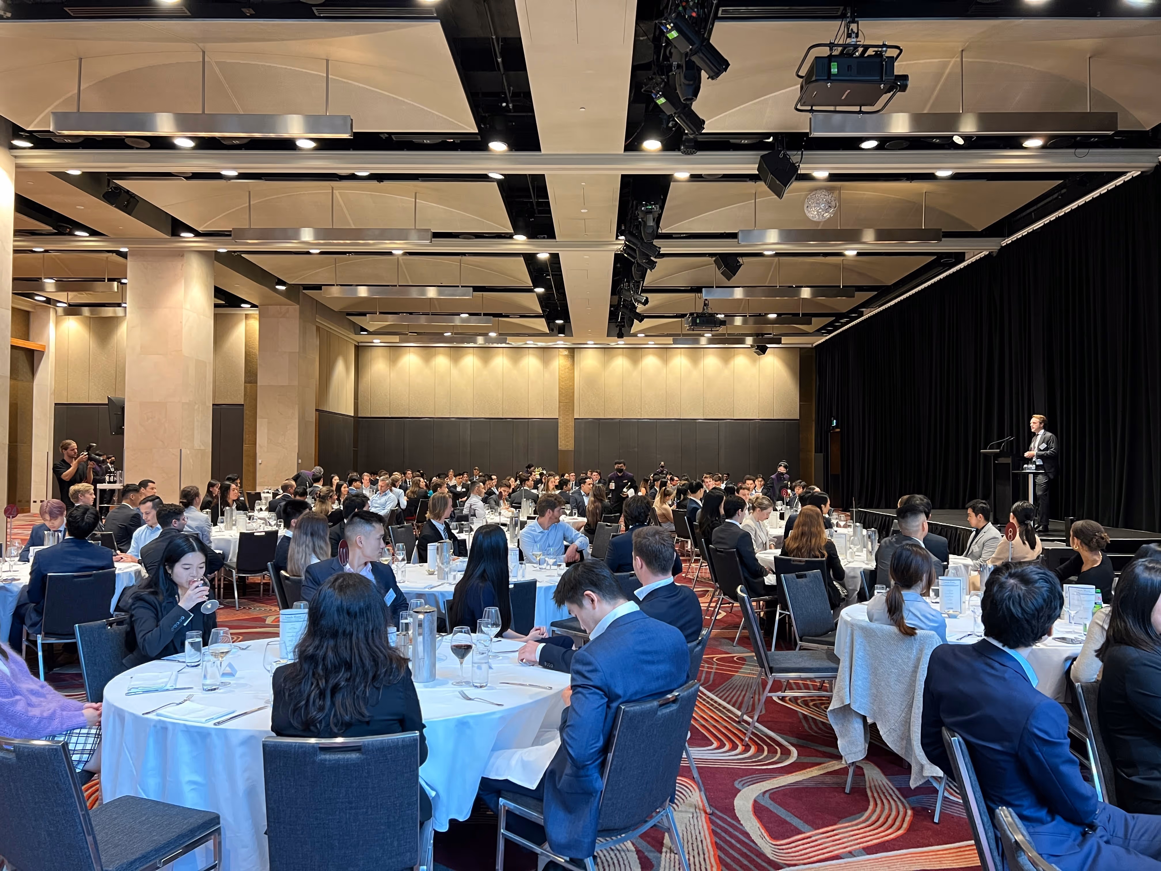 Audience dressed in business attire seated at round tables listening to a speaker on stage in a conference room.