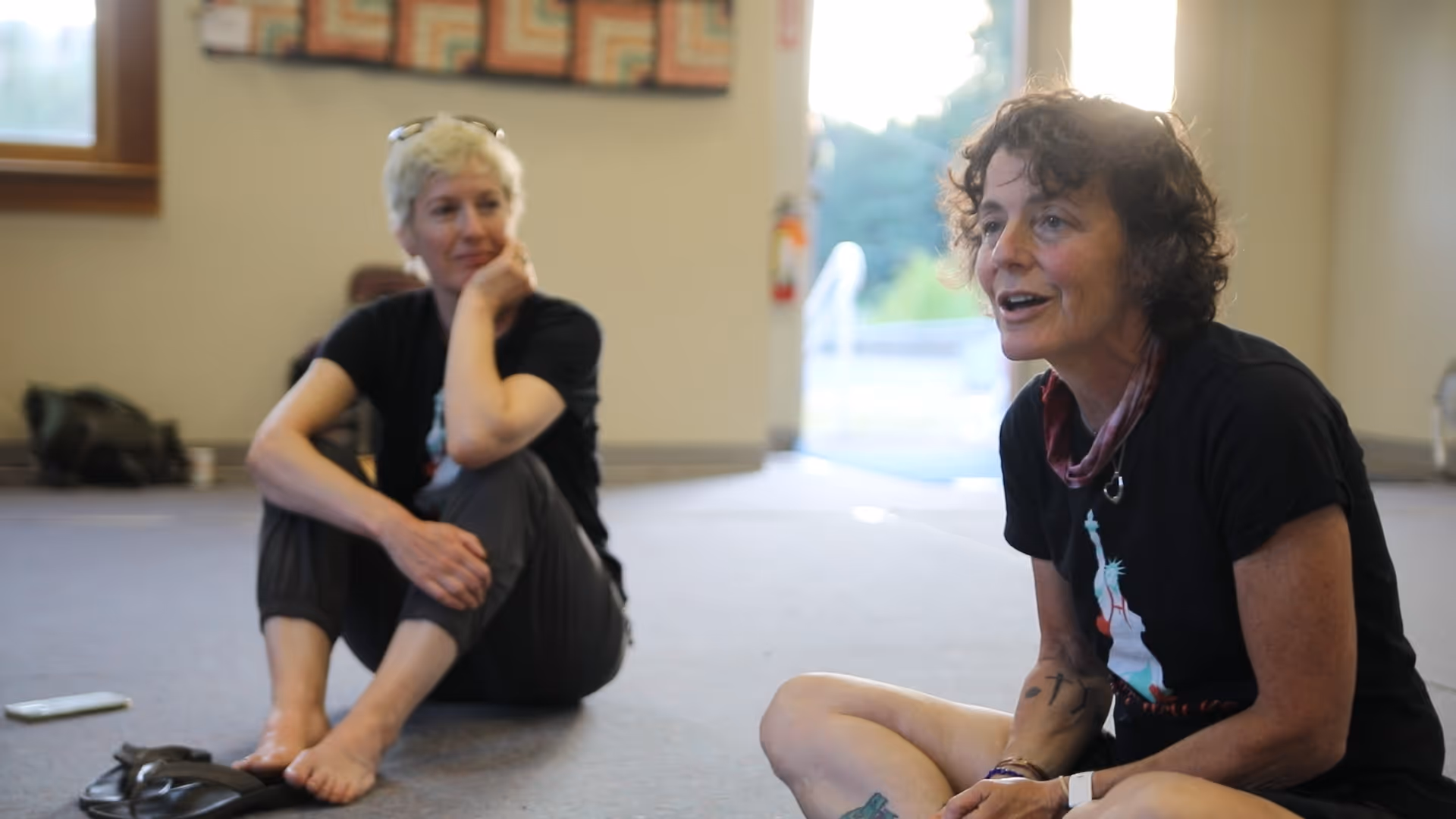 Two women sitting on the floor indoors, one speaking and the other listening attentively.