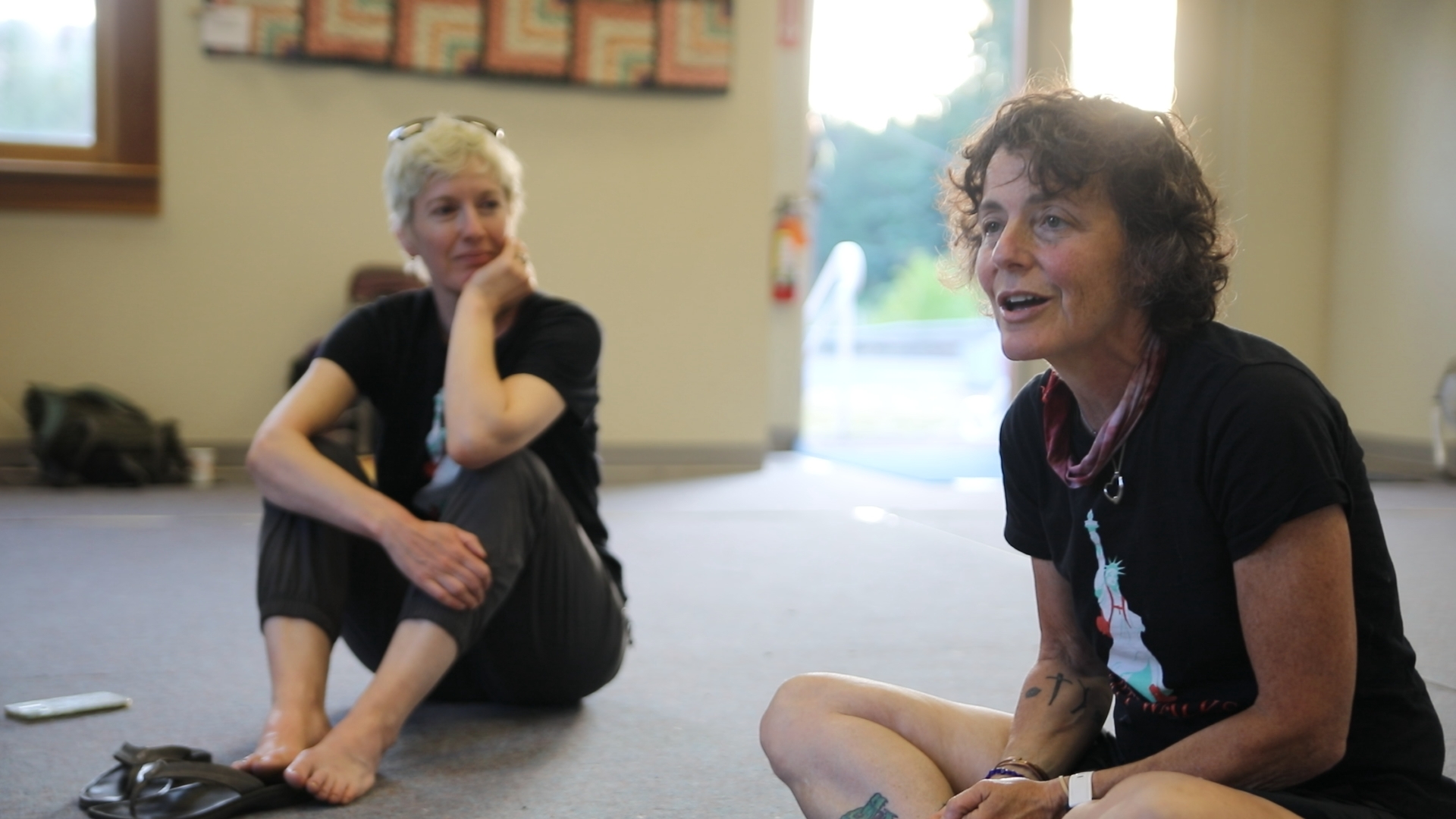 Two women sitting on the floor indoors, one speaking and the other listening attentively.