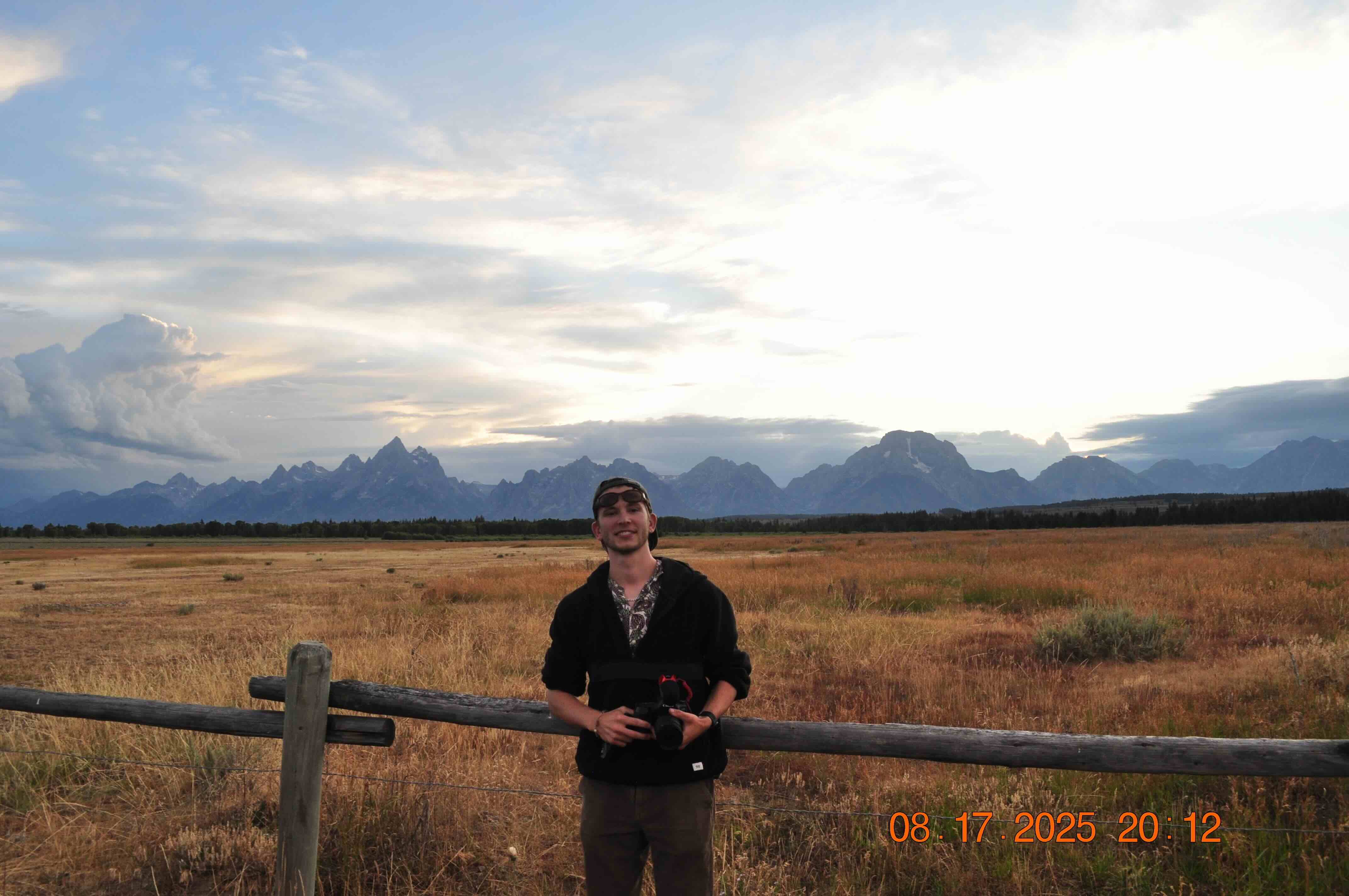 A young man holding a camera stands in front of a wooden fence with a vast dry grass field and mountain range under a cloudy sky at sunset.