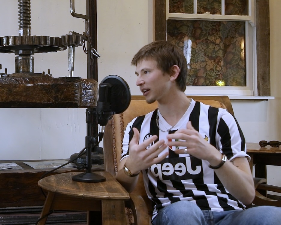 Young man in a black and white striped soccer jersey speaking into a microphone while gesturing with his hands indoors.