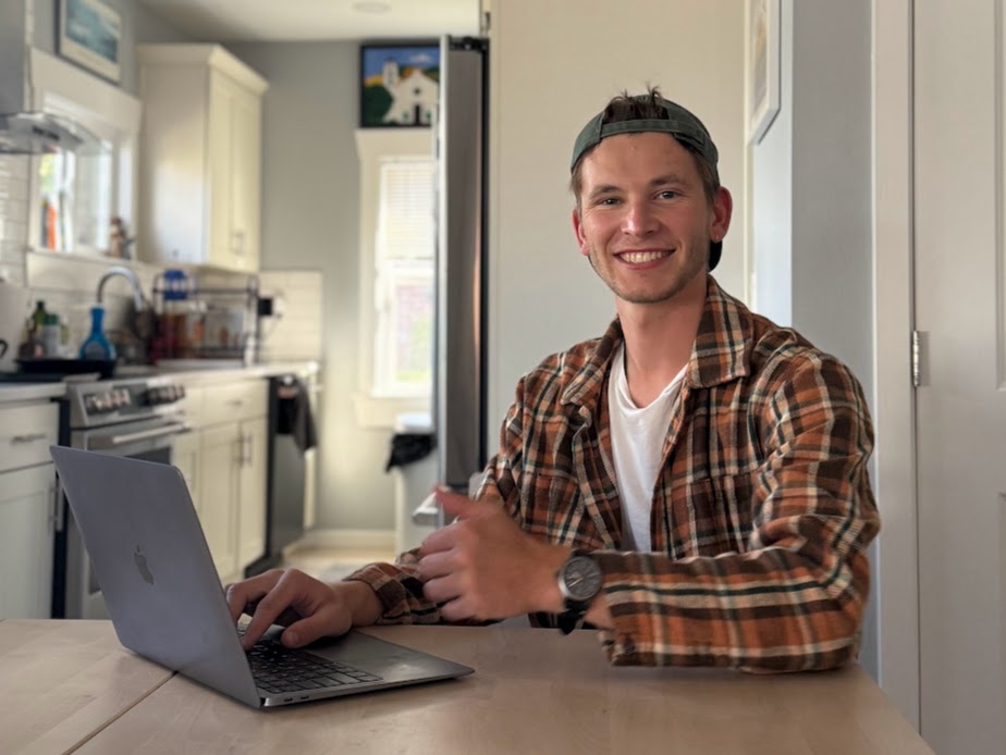 Smiling young man wearing a backward cap and plaid shirt working on a laptop at a wooden table in a bright kitchen.