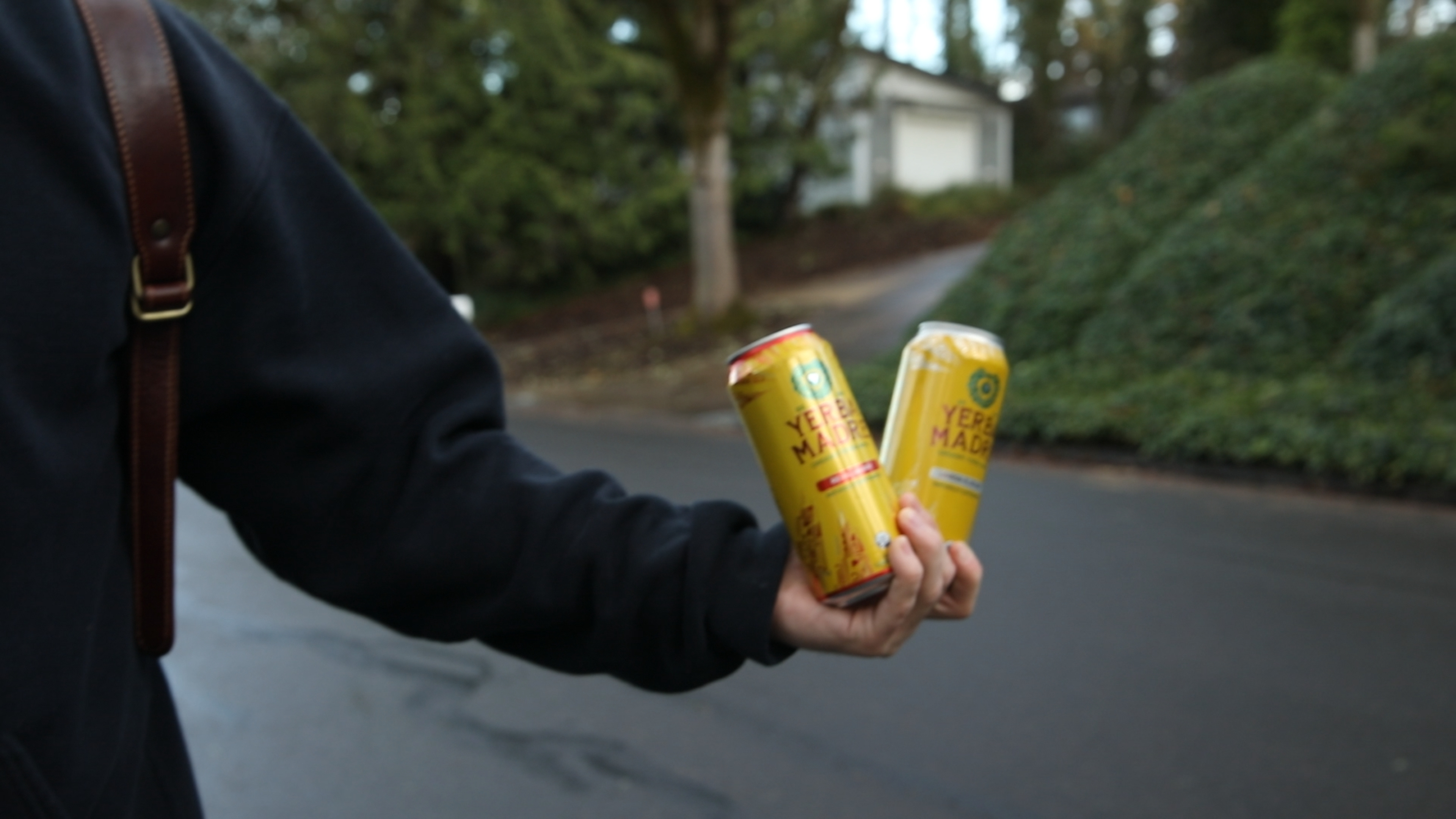 Person holding two yellow cans of Yerbsi Made beverage on a street with greenery and houses in the background.