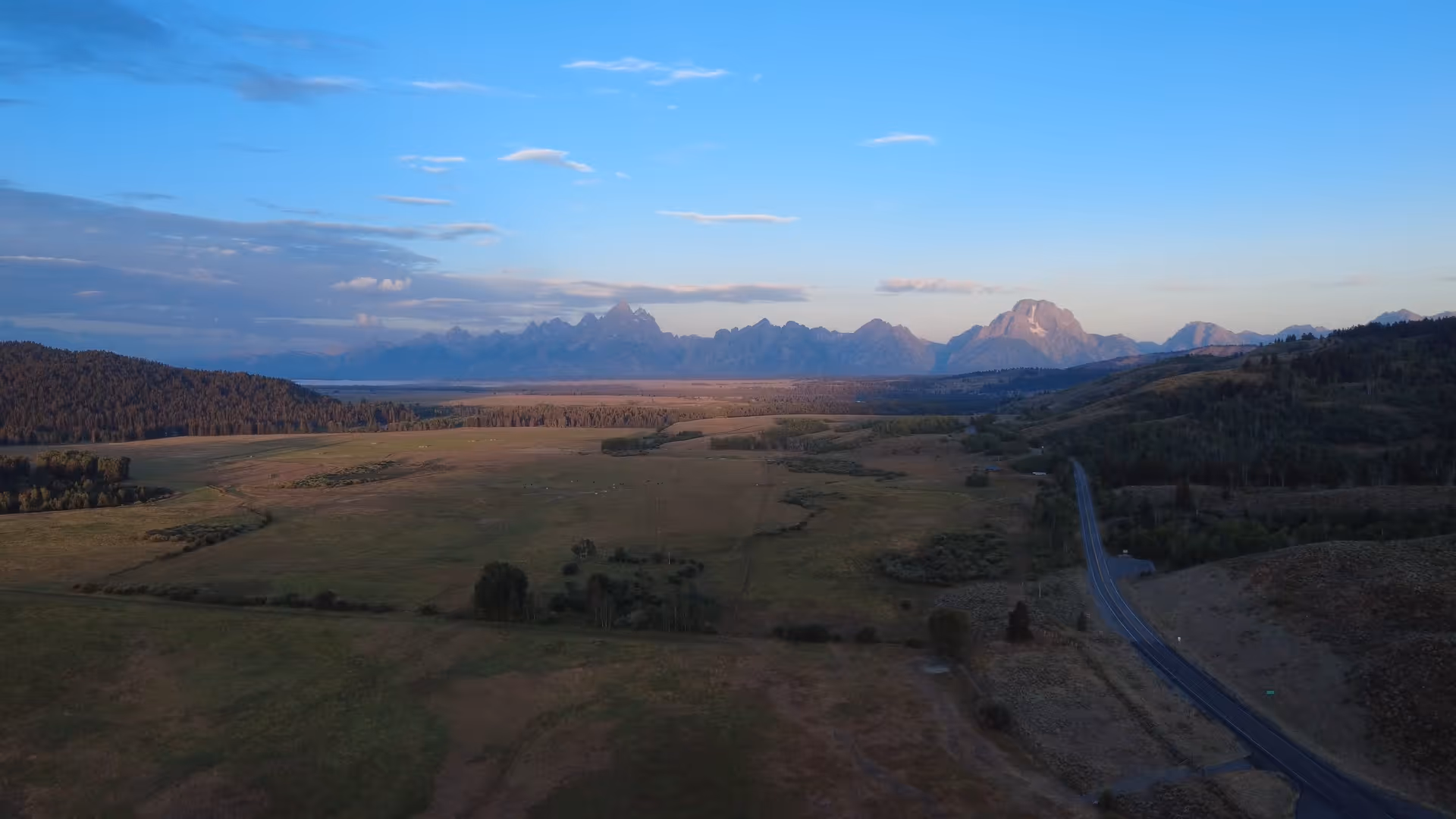 Aerial view of a valley with patchy fields, a winding road on the right, and a mountain range under a blue sky with scattered clouds at sunset.