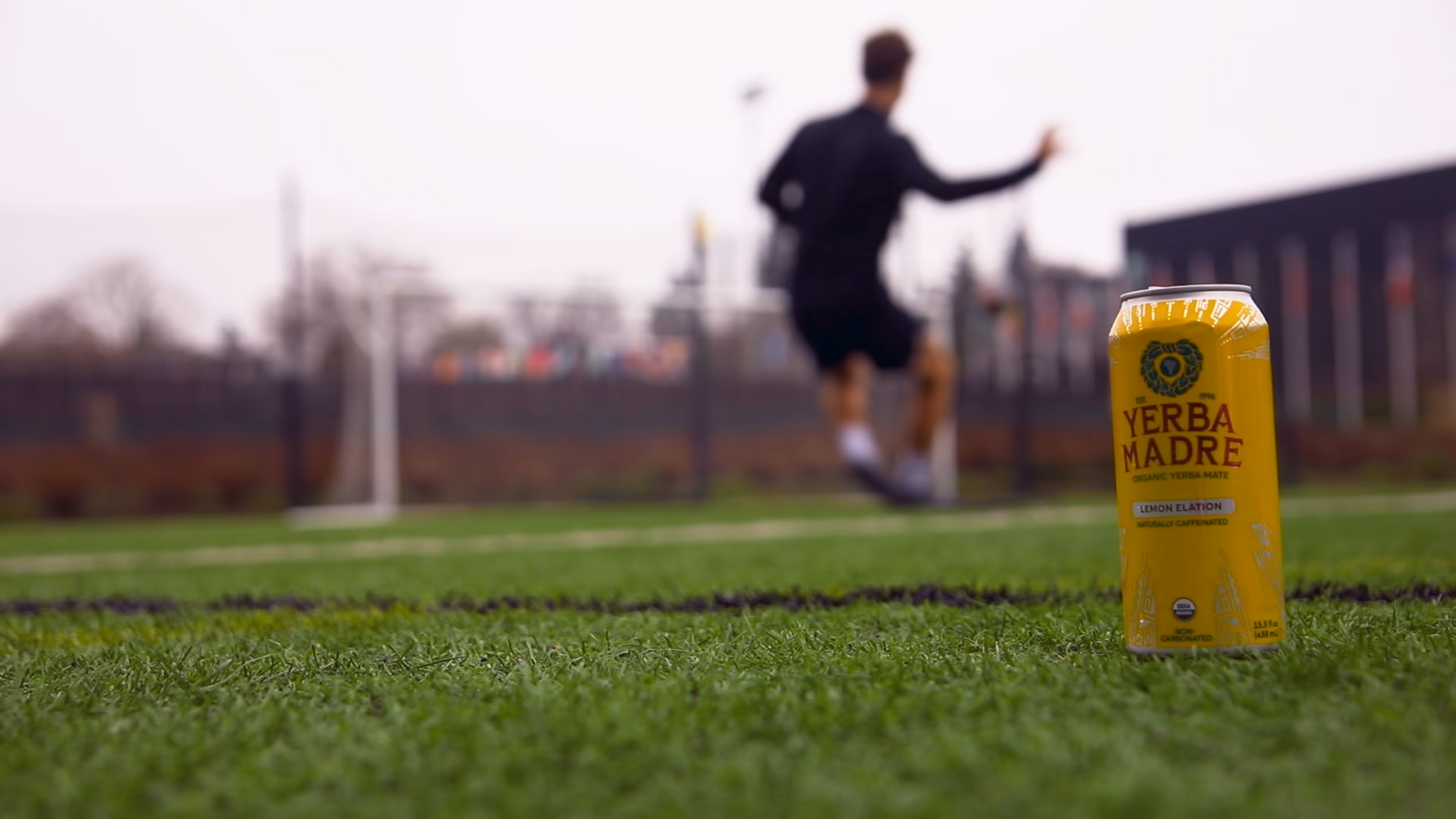 Yellow can of Yerba Madre Lemon Elation yerba mate on grass with blurred soccer player in background.
