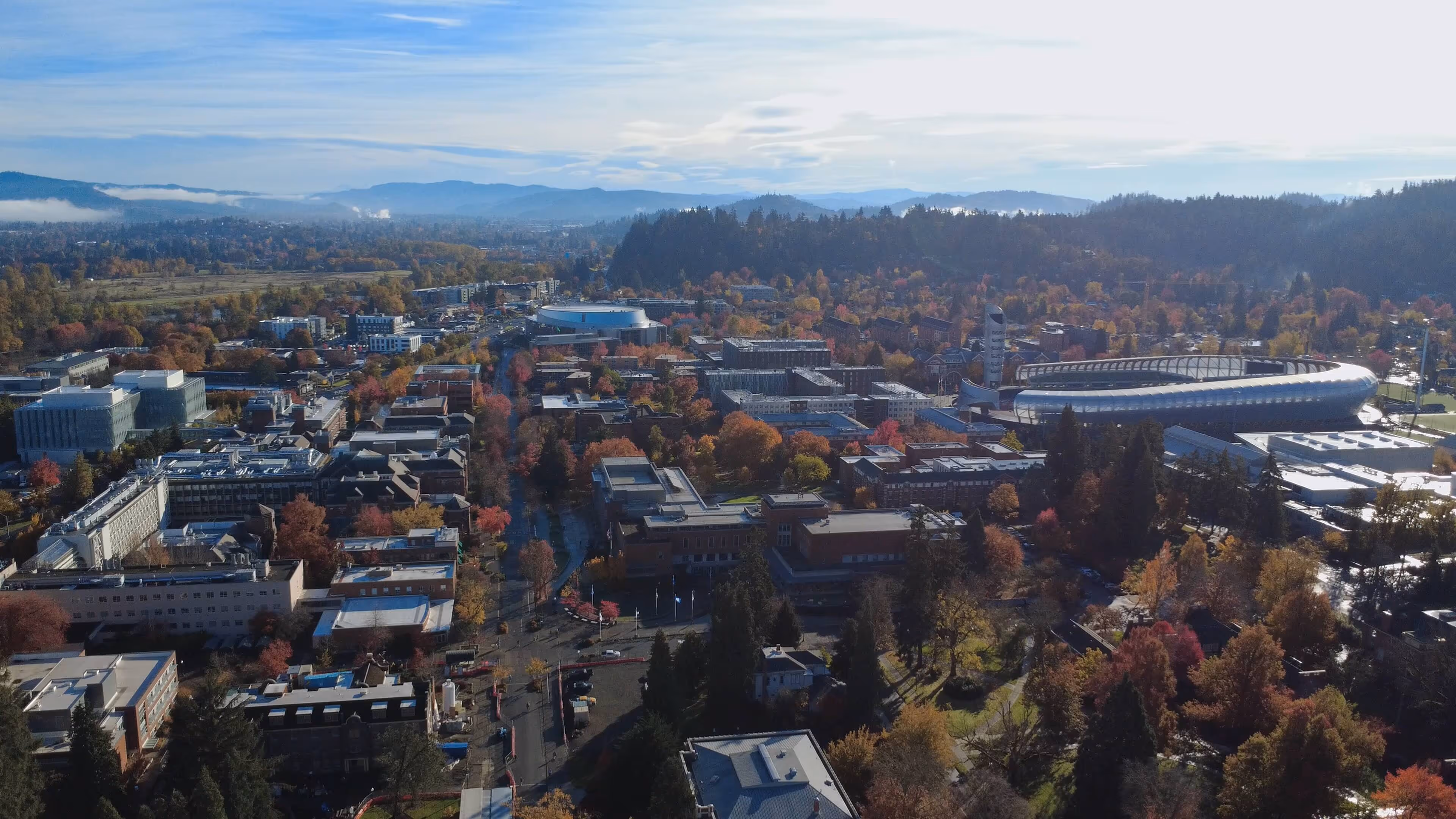 Aerial view of a college campus with modern buildings, a circular stadium, autumn-colored trees, and mountains in the background under a partly cloudy sky.