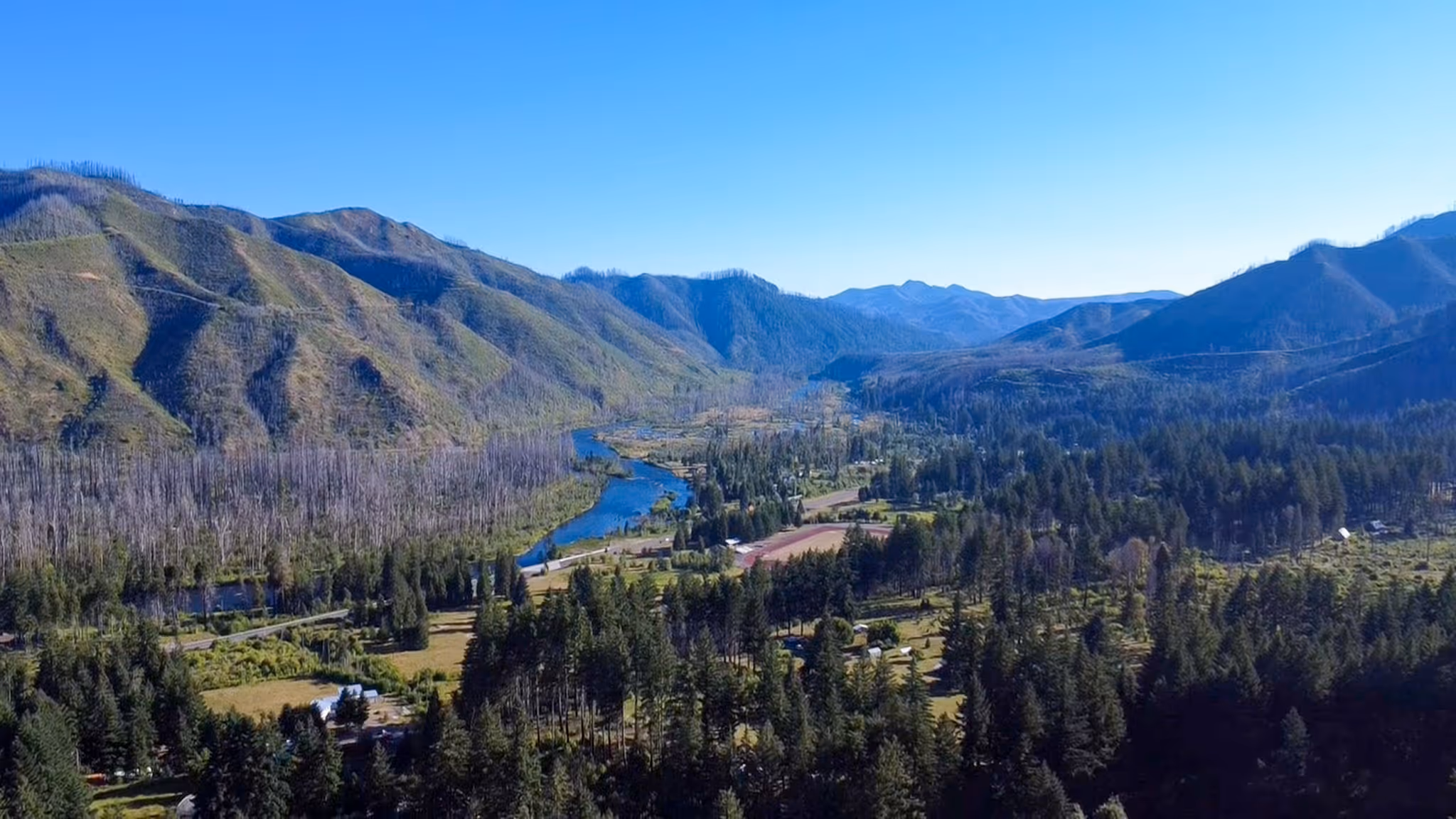 A winding river flows through a valley surrounded by forested mountains under a clear blue sky.