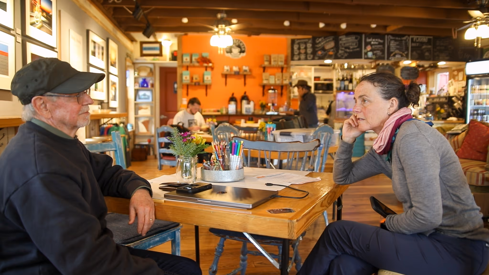 Two people sitting and talking across a wooden table in a cozy cafe with art on the walls and other patrons in the background.