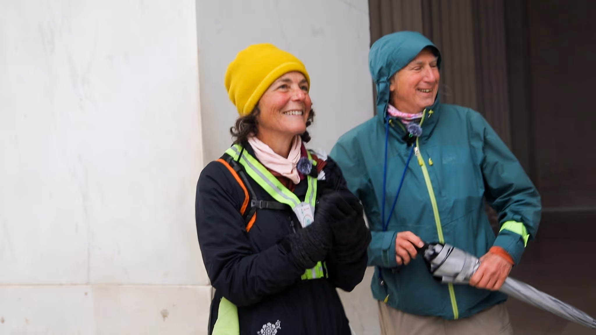 Two smiling people dressed in outdoor jackets, one wearing a yellow beanie and the other holding a folded umbrella.