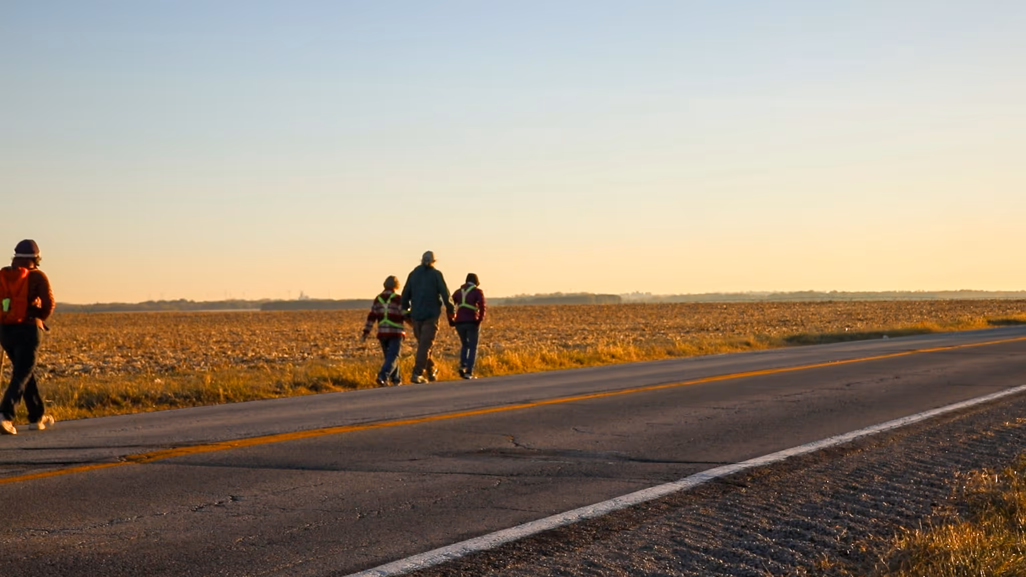 Three people walking along a rural road beside a vast field during sunset with one person trailing behind.