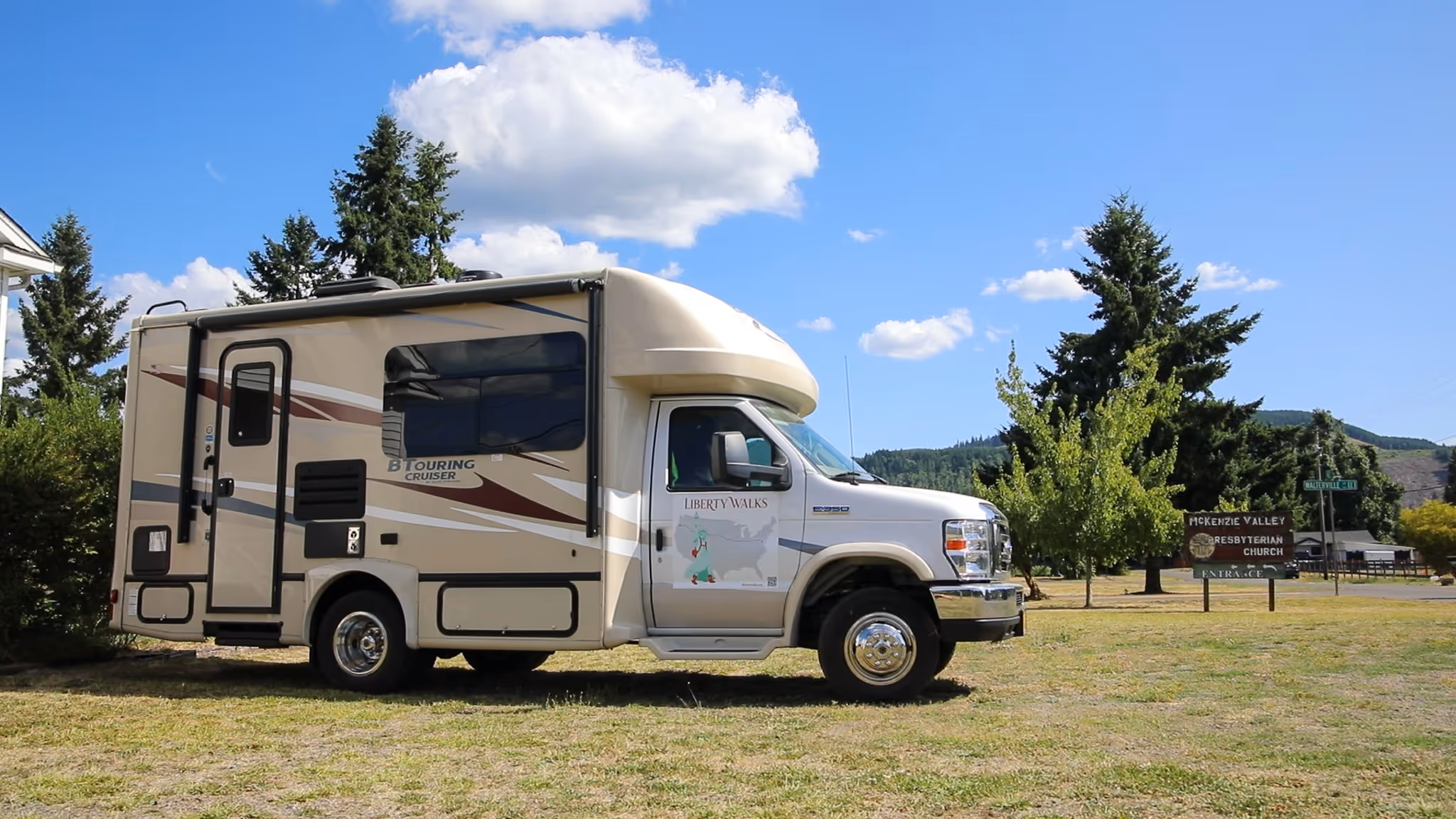 Beige B Touring Cruiser RV parked on grass near McKenzie Valley Presbyterian Church sign and trees under a blue sky with clouds.