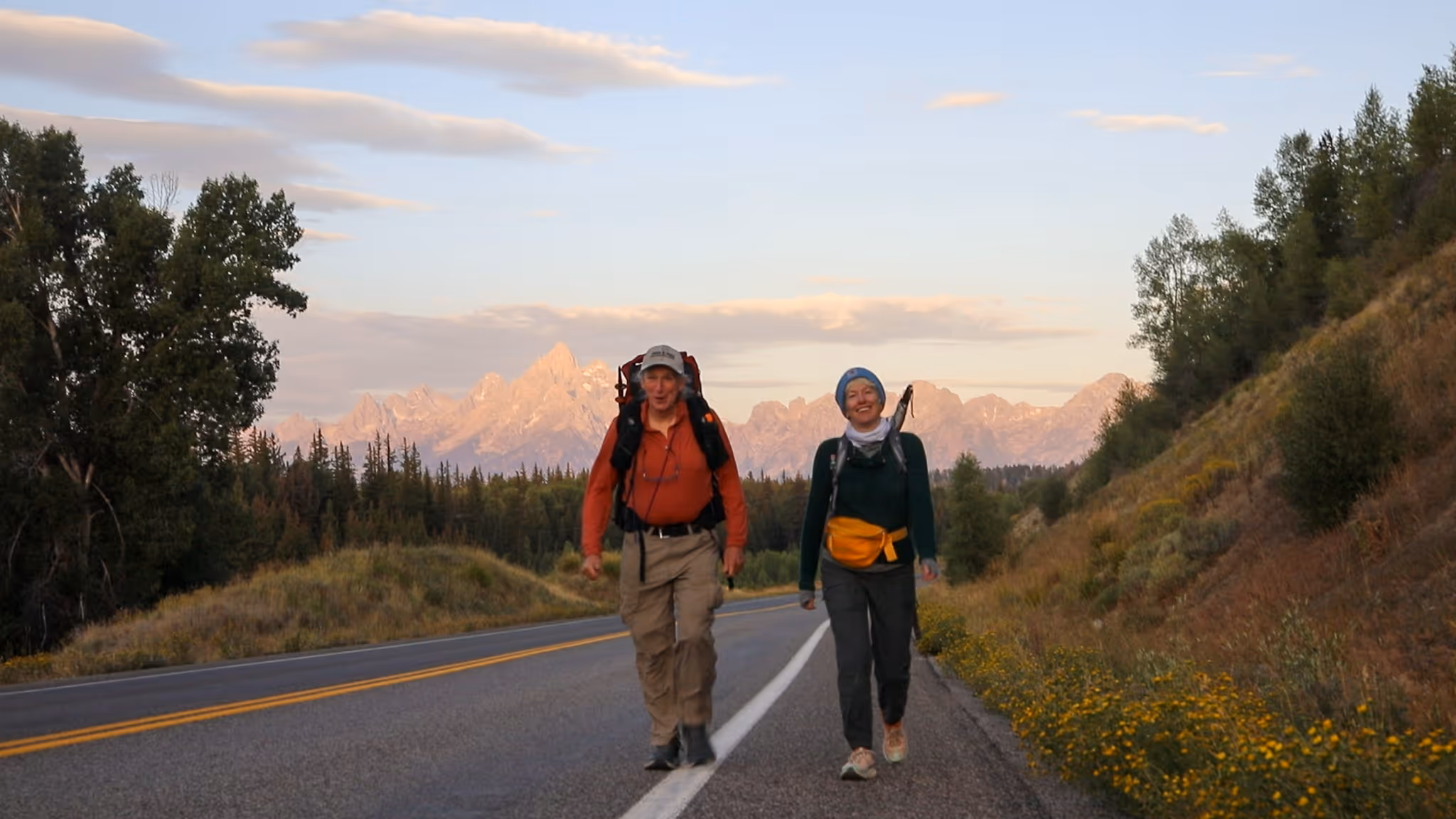 Two women embracing warmly outdoors with a group of smiling people watching behind them.