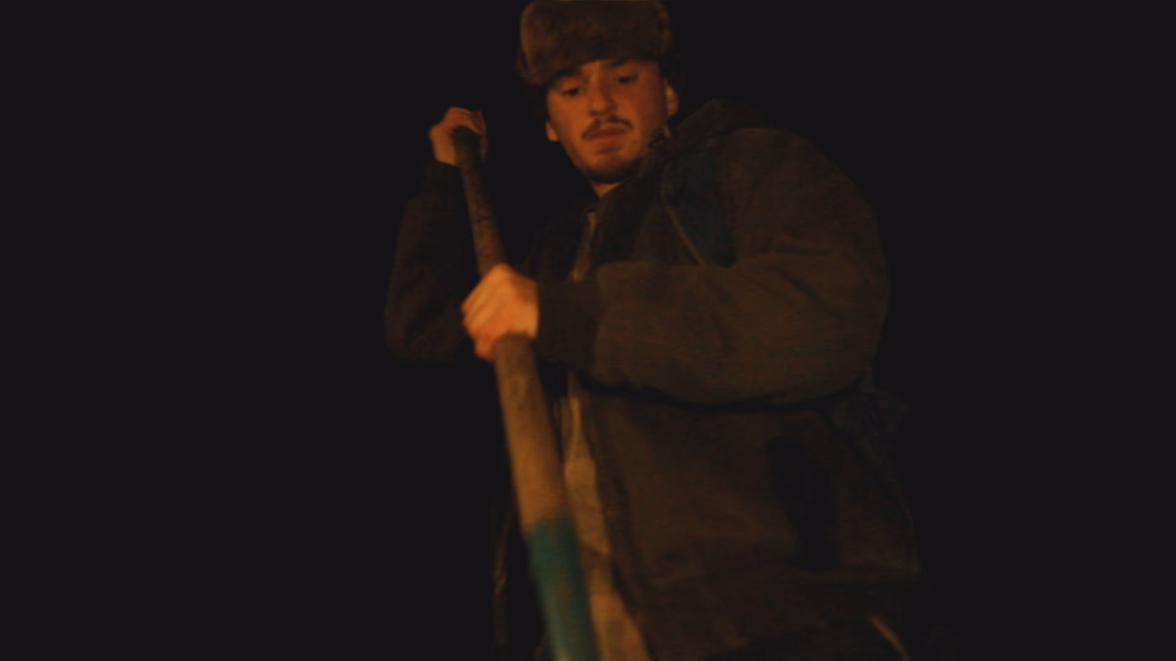 Young man wearing a furry hat and dark jacket holding a wooden pole against a dark background.